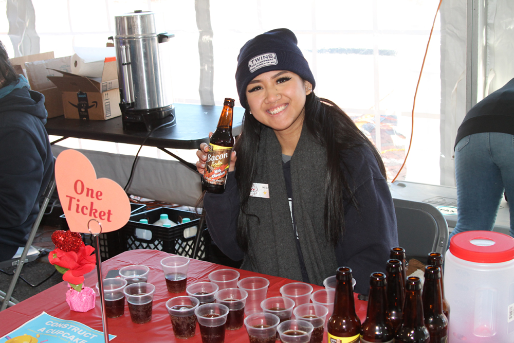 woman wearing a beanie with a patch that reads "Twins Ace Hardware" is holding a bottle of bacon soda (yes, bacon soda) selling tickets at the Chocolate Lovers Festival
